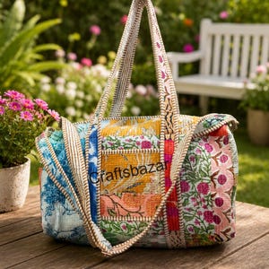 Könnte beinhalten: Eine rechteckige Reisetasche aus gestepptem Stoff mit einem Patchwork aus Blumen- und geometrischen Mustern in Blau, Gelb, Rosa und Grün. Die Tasche verfügt über zwei lange Schulterriemen und den Schriftzug Craftsbazar auf der Vorderseite.