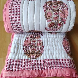 Könnte beinhalten: Eine gefaltete, gesteppte Decke mit floralem Design. Die Decke hat einen weißen Hintergrund mit rosa und braunen Blumenmustern. Die Ränder sind mit einem einfarbigen rosa Rand und einem gemusterten Band aus Weiß und Rosa versehen. Die Decke ist aus weichem, gestepptem Stoff.