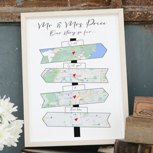 May include: A white framed print with a map-themed design. The print features a series of directional signs with maps and red hearts. The text on the signs reads "We met", "Will you?", "I +", and "Our home". The print is titled "Mr & Mrs Price Our story so far..."