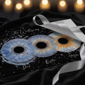 May include: A personalised art print with three iris eye designs in blue and gold, with names and the family name. The print is on a black background and is decorated with a silver ribbon.