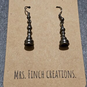 May include: Silver-coloured earrings in the shape of chess pieces, presented on a brown card. The card features the text "MRS. FINCH CREATIONS." The earrings have a detailed, decorative design.