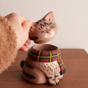 Könnte beinhalten: Ein Keramikgefäß in Katzenform mit abnehmbarem Deckel. Die Katze ist braun mit dunklen Streifen und trägt einen rot-schwarz-gelben Schal mit Karomuster. Der Deckel zeigt ein gemaltes Katzengesicht mit einer Brille aus goldenem Draht.