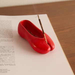 May include: A vibrant red shoe-shaped incense holder with a brown incense stick. The holder sits on a white page with printed text. The shoe features a split toe design.