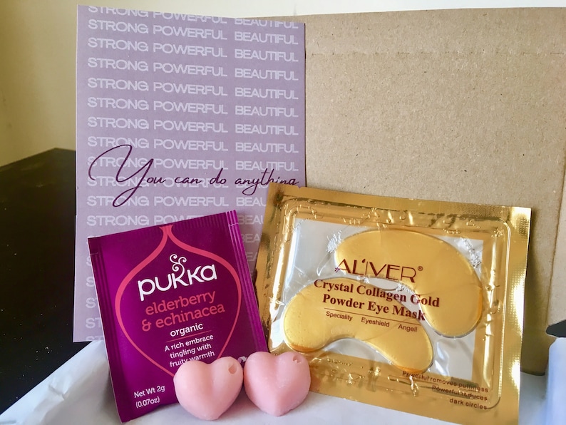 May include: A purple tea bag with the text "pukka elderberry & echinacea organic" and a pink heart-shaped bath bomb. A gold packet with the text "ALIVER Crystal Collagen Gold Powder Eye Mask" and a pink heart-shaped bath bomb. A purple card with the text "STRONG POWERFUL BEAUTIFUL" repeated multiple times and the text "You can do anything".