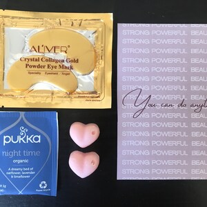 May include: A set of self-care items including a pink heart-shaped bath bomb, a blue tea bag with the text "pukka night time organic", a gold packet of eye masks with the text "ALIVER Crystal Collagen Gold Powder Eye Mask", and a purple card with the text "STRONG POWERFUL BEAUTIFUL" repeated multiple times and the text "You can do anything" in the center.