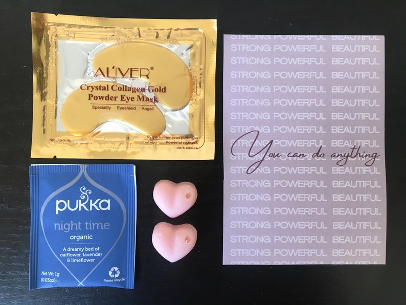 May include: A set of self-care items including a pink heart-shaped bath bomb, a blue tea bag with the text "pukka night time organic", a gold packet of eye masks with the text "ALIVER Crystal Collagen Gold Powder Eye Mask", and a purple card with the text "STRONG POWERFUL BEAUTIFUL" repeated multiple times and the text "You can do anything" in the center.