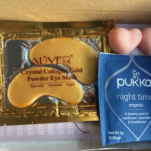 May include: A close-up of a box containing three beauty products: a gold packet of ALIVER Crystal Collagen Gold Powder Eye Mask, two pink heart-shaped bath bombs, and a blue packet of PUKKA night time organic tea. The tea packet has the text "A dreamy bed of oatflower, lavender & limeflower" and "Net Wt 1g (0.03oz)".
