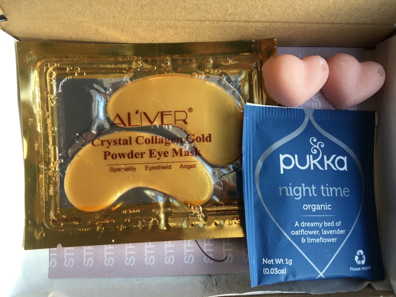 May include: A close-up of a box containing three beauty products: a gold packet of ALIVER Crystal Collagen Gold Powder Eye Mask, two pink heart-shaped bath bombs, and a blue packet of PUKKA night time organic tea. The tea packet has the text "A dreamy bed of oatflower, lavender & limeflower" and "Net Wt 1g (0.03oz)".
