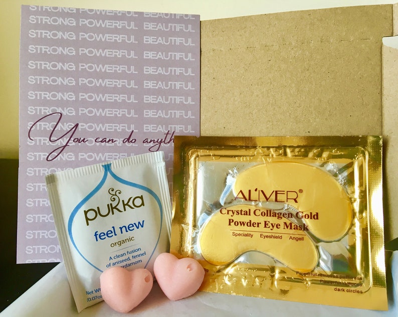 May include: A close-up of a small cardboard box with a pink card that says "You can do anything" and a gold pouch with the text "ALIVER Crystal Collagen Gold Powder Eye Mask" and two pink heart-shaped bath bombs. The pouch also has the text "Speciality Eyeshield Angell" and "dark circles". There is also a small white pouch with the text "pukka feel new organic" and "A clean fusion of aniseed, fennel cardamom".