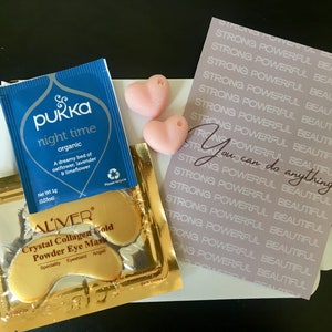 May include: A flat lay of self-care items including a blue tea bag with the text "Pukka night time organic", two pink heart-shaped soaps, a gold packet of eye masks with the text "ALIVER Crystal Collagen Gold Powder Eye Mask", and a pink card with the text "STRONG POWERFUL BEAUTIFUL" repeated multiple times and the text "You can do anything" in a larger font.