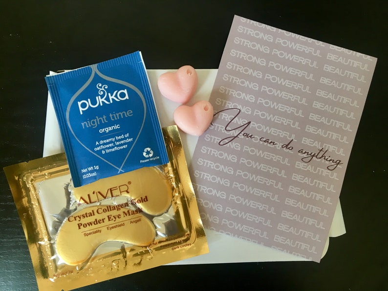 May include: A flat lay of self-care items including a blue tea bag with the text "Pukka night time organic", two pink heart-shaped soaps, a gold packet of eye masks with the text "ALIVER Crystal Collagen Gold Powder Eye Mask", and a pink card with the text "STRONG POWERFUL BEAUTIFUL" repeated multiple times and the text "You can do anything" in a larger font.