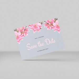以下が含まれることがあります： ピンクの花のアクセントが付いた薄い青色の結婚式の招待状。テキストは「Mia + Stephen Save the Date 2025年2月14日」です。