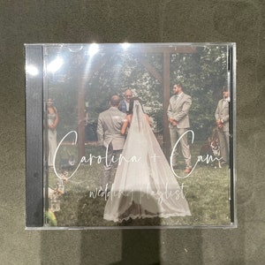 May include: A wedding CD case with a photo of the bride and groom in front of a wooden archway. The text "Carolina + Cam Wedding Playlist" is written in white cursive on the front of the case.