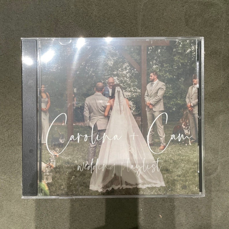 May include: A wedding CD case with a photo of the bride and groom in front of a wooden archway. The text "Carolina + Cam Wedding Playlist" is written in white cursive on the front of the case.
