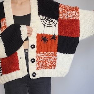 Puede incluir: Cárdigan grueso de patchwork en crema, negro y naranja quemado. El suéter presenta un diseño de telaraña y arañas en la parte delantera. Tiene botones negros grandes y mangas anchas. El estilo general es un ajuste cómodo y de gran tamaño.