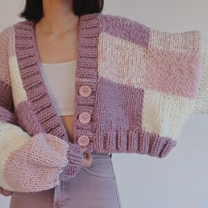 Op de afbeelding: Een gecropt, dik gebreid vest in tinten lavendel, roze en crème. De trui heeft een patchwork-ontwerp met grote, vierkante kleurblokken en oversized knopen. De mouwen zijn wijd en de algehele stijl is gezellig.