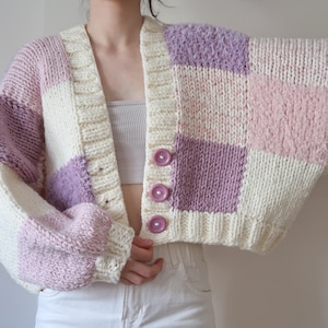 May include: A cream, pink, and lavender patchwork cardigan. The chunky knit design features large squares of colour. It has a V-neck, three pink buttons, and wide sleeves.