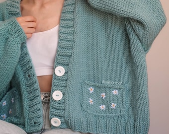 May include: A light blue knitted cardigan with white buttons and a pocket on the front. The pocket has embroidered white daisies. #teesooda