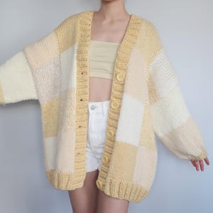 May include: An oversized, chunky cardigan with a patchwork design. The sweater features large, square panels in shades of yellow and cream, with large, round buttons. The sleeves are long and the fit is relaxed, a cozy knitwear item.
