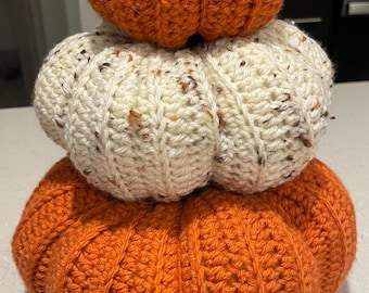 May include: A stack of three crocheted pumpkins in shades of orange and cream. The bottom and top pumpkins are orange, while the middle one is cream with flecks of brown. The pumpkins are stacked on top of each other.