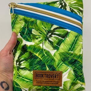 May include: Quilted book sleeve with a vibrant green and white tropical leaf pattern. Features a blue and white accent stripe with a gold zipper. A brown leather patch reads "BOOKTROVERT".