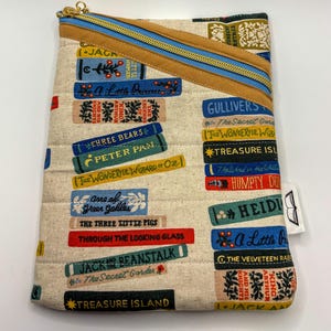May include: A beige quilted book-themed pouch with a gold zipper and blue accents. The pouch features colorful book titles like "Treasure Island" and "Peter Pan". A small white tag with a book icon is attached.