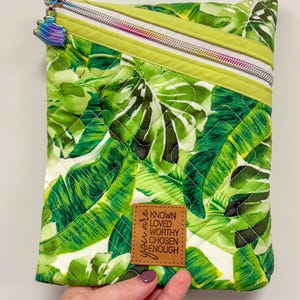 May include: A quilted pouch with a vibrant green tropical leaf pattern. The pouch features a diagonal zipper with a rainbow-colored pull and a small brown leather tag with the words "You are Known, Loved, Worthy, Chosen, Enough."