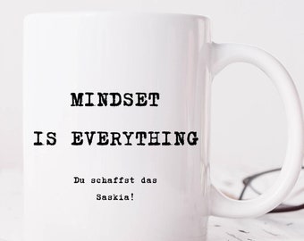 以下が含まれることがあります： 白地に黒い文字で「MINDSET IS EVERYTHING」と「Du schaffst das Saskia!」と書かれた白いセラミック製のマグカップです。