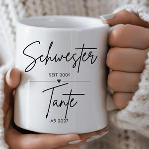 Puede incluir: Una taza de cerámica blanca con texto impreso en negro que dice "Schwester SEIT 2001" sobre un pequeño icono de corazón y "Tante AB 2027" debajo. La taza tiene un cuerpo cilíndrico liso con un asa en el lado derecho.