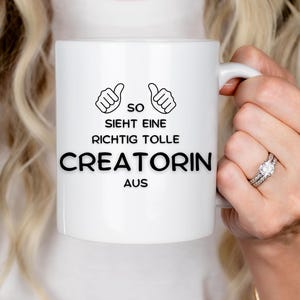 Könnte beinhalten: Weißer Keramikbecher mit dem Text "so sieht eine richtig tolle CREATORIN aus" in Schwarz. Der Becher zeigt zwei Daumen hoch Illustrationen über dem Text. Der Becher wird von einer Person gehalten.