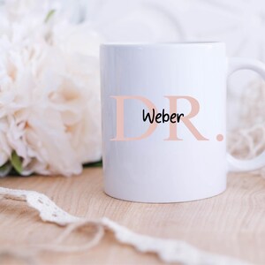 Peut inclure: Une tasse en céramique blanche avec un motif rose "DR. Weber". La tasse a une anse et est posée sur une surface en bois.