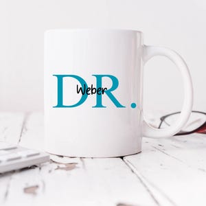 Pode incluir: Caneca de cerâmica branca com um design azul "DR." e o nome "Weber" escrito em itálico dentro do "D".