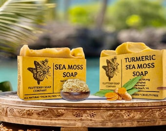 May include: Two rectangular bars of soap with tan paper labels. The left label reads Sea Moss Soap, Flutterby Soap Company. The right label reads Turmeric Sea Moss Soap, Flutterby Soap Company. A small wooden bowl of sea moss and turmeric root sit in front.