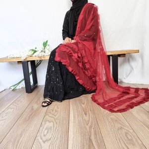 May include: A black sequined dress with a burgundy tulle scarf. The scarf has a ruffled edge and is decorated with small, sparkling details. The outfit is finished with black sandals.