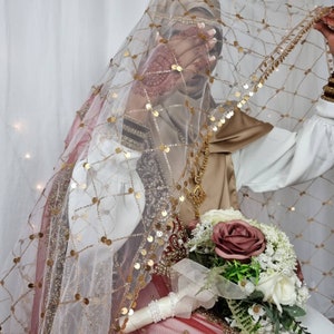 May include: A bride wearing a white and gold embroidered veil with a red and gold embroidered dress. She is holding a bouquet of pink and white flowers.