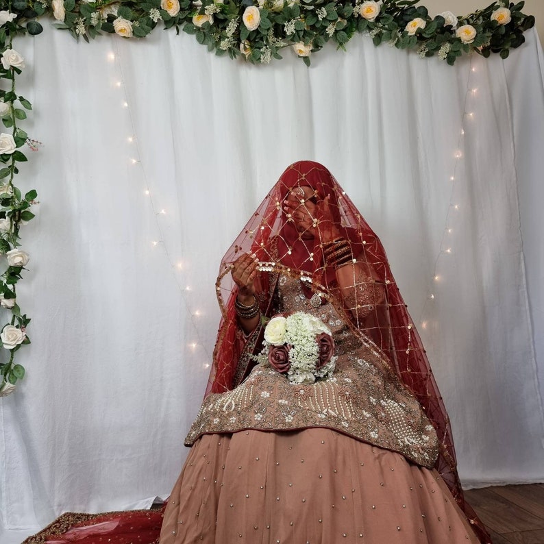 May include: A bride wearing a red and gold embroidered dress with a red veil. She is holding a bouquet of white and pink flowers.