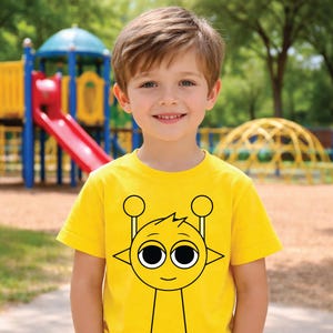 Könnte beinhalten: Ein gelbes T-Shirt mit einem Cartoon-Alien-Design, getragen von einem Kind. Das Alien hat große Augen und Antennen. Im Hintergrund ist ein Spielplatz mit Rutschen und Klettergerüsten zu sehen.