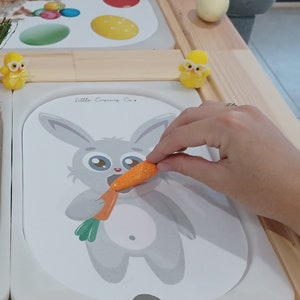 May include: A white ceramic dish filled with a nest of straw and speckled Easter eggs.  There are small yellow chicks and orange carrots scattered throughout the nest.  A white ceramic dish with a gray bunny illustration and the text "Little Bunny Fun" is next to the nest.