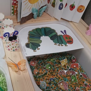 Op de afbeelding: Een houten speeltafel met vier containers. De containers zijn gevuld met groen en bruin versnipperd papier, groene en bruine linzen en een verscheidenheid aan kleurrijke viltvoedsel. Er zijn ook verschillende kleurrijke viltvlinders en een rups-illustratie.