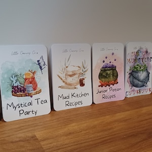 May include: A collection of rectangular cards with rounded corners, featuring watercolor illustrations and text. The cards include titles like "Mystical Tea Party," "Mud Kitchen Recipes," and "Junior Potion Recipes." The cards are arranged on a wooden surface.