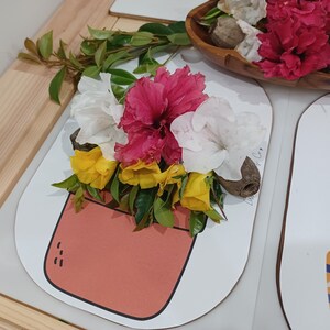 Puede incluir: Arreglo floral con flores rosas, blancas y amarillas sobre una tarjeta blanca con una ilustración de un jarrón color melocotón. Hojas verdes y una vaina de madera añaden elementos naturales. Un cuenco de madera con flores está en el fondo.