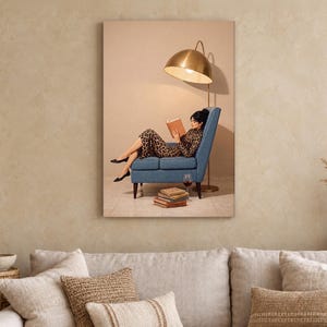 May include: A framed wall art print depicting a person in a leopard print dress sitting in a blue armchair, reading a book under a large gold dome floor lamp. A stack of books and a glass of red wine are placed on the floor near the chair.