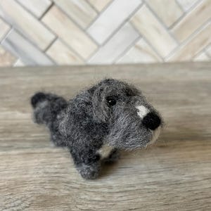 May include: A gray and white needle felted dog figurine. The dog is sitting up with its head tilted up and its tongue sticking out.