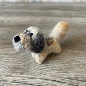 May include: A small, hand-felted dog figurine with a brown, white, and black wool blend. The dog has a playful expression with black eyes and a brown nose.