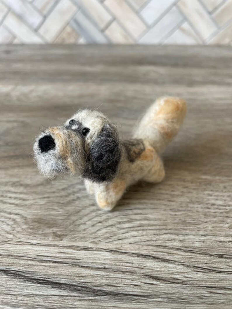 May include: A small, hand-felted dog figurine with a brown, white, and black wool blend. The dog has a playful expression with black eyes and a brown nose.