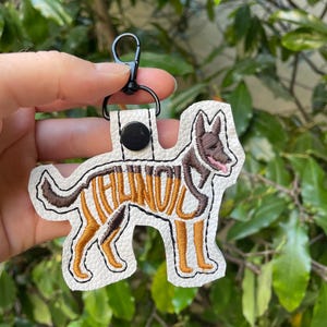 May include: A white leather keychain featuring a dog design with brown and tan embroidery. The word "ILLINOIS" is embroidered on the dog's body. The keychain has a black clasp and a black button.