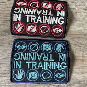 May include: Two rectangular embroidered patches with black backgrounds. The top patch features white text reading IN TRAINING with red prohibition symbols over icons of an eye, speech bubble, camera, and hand. The bottom patch has blue text and icons.