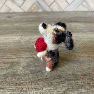 May include: A small, felted dog figurine holding a red heart. The dog is black, white, and brown with a black nose and black eyes.