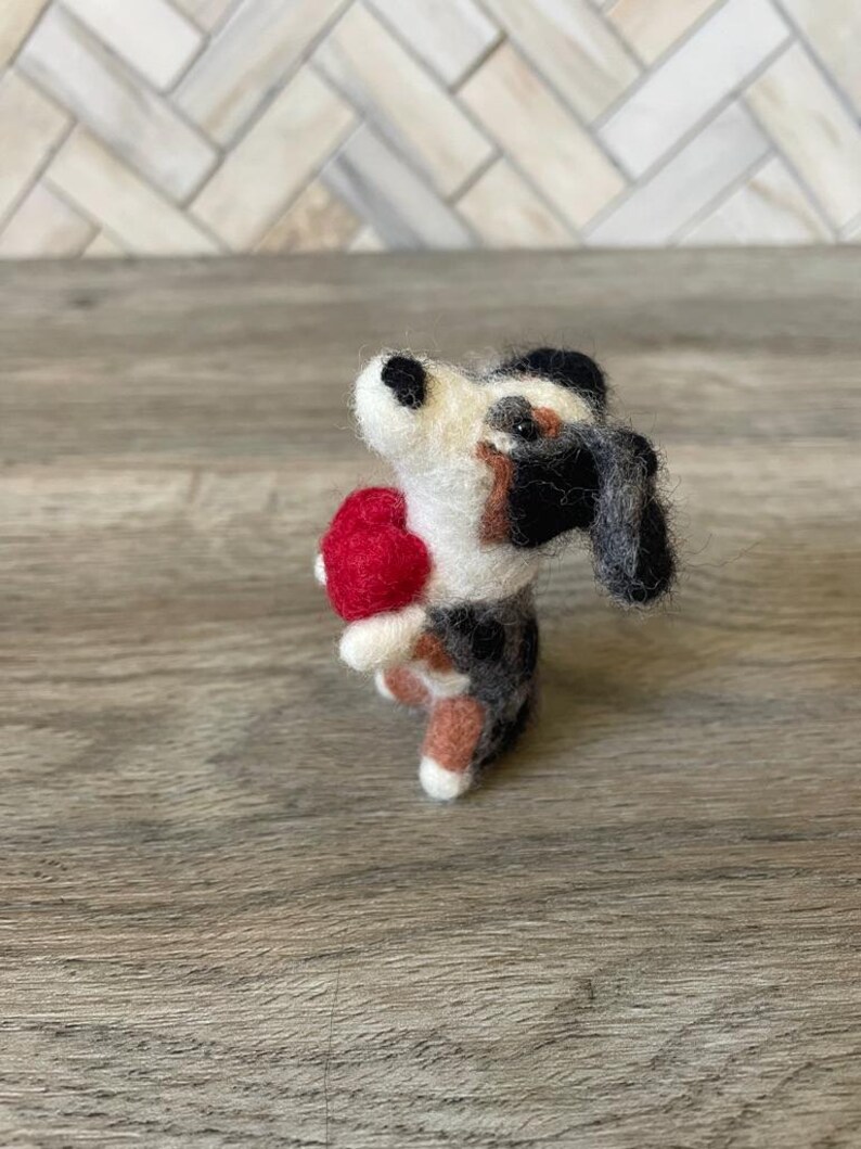 May include: A small, felted dog figurine holding a red heart. The dog is black, white, and brown with a black nose and black eyes.