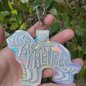 Puede incluir: Un llavero con forma de perro Gran Pirineo con material holográfico iridiscente. Las palabras "GREAT PYRENEES" están bordadas con hilo plateado. El llavero tiene un cierre plateado.